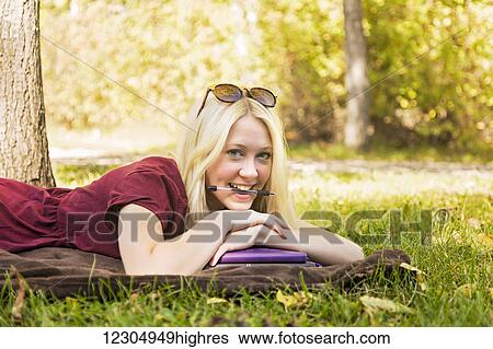Stock Photograph - A young woman doing homework outdoors while enjoying a warm autumn day in a park and posing for the camera with a pen in her mouth; Edmonton, Alberta, Canada. Fotosearch