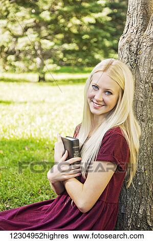 Stock Photograph - A young woman holding her Bible close to her chest while spending personal devotional time outdoors in a park in autumn; Edmonton, Alberta, Canada. Fotosearch
