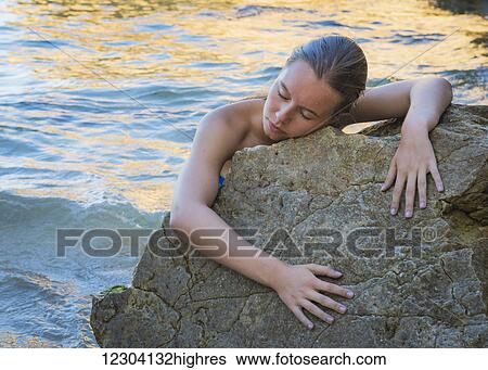 A young woman in a bikini clings to a rock at the water's edge with her eyes closed; Faro, Portugal View Large Photo Image Stock Photograph - A young woman in a bikini clings to a rock at the water's edge with her eyes closed; Faro, Portugal. Fotosearch