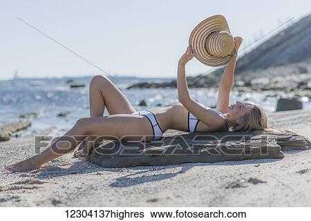 A young woman in a bikini lays on a piece of driftwood on the beach holding a sunhat up to shield the sun; Tarifa, Cadiz, Andalusia, Spain View Large Photo Image Stock Photograph - A young woman in a bikini lays on a piece of driftwood on the beach holding a sunhat up to shield the sun; Tarifa, Cadiz, Andalusia, Spain. Fotosearch