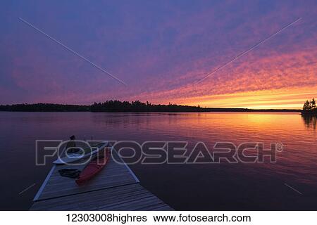 Stock Photograph - A young woman sits at the end of the dock with kayaks watching the sun set over a lake; Ontario, Canada. Fotosearch