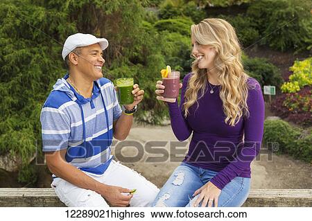 Stock Photograph - An adult couple looks at each other smiling and toasting one another with fresh fruit smoothies in a picnic park setting; Vancouver, British Columbia, Canada. Fotosearch