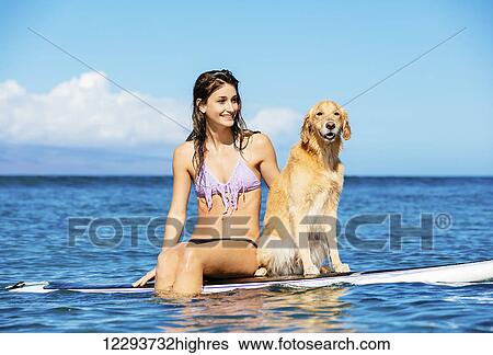 Stock Photograph - Attractive Young Woman Surfing with her Dog. Sharing surfboard with Golden Retriever.. Fotosearch