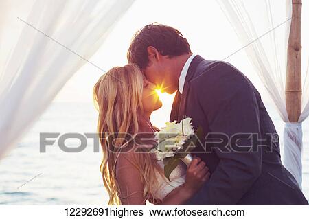 Stock Photograph - Beautiful Sunset Wedding by the Sea. Bride and Groom Kissing at Sunset. Romantic Married Couple.. Fotosearch