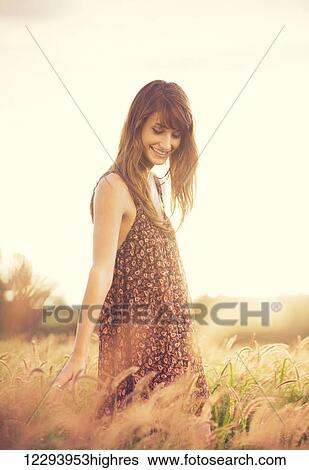 Stock Photograph - Beautiful young woman outdoors. Romantic Model in Sun Dress in Golden Field at Sunset. Happy Emotions, Glowing Sunlight. Backlit. Warm color tones. Fotosearch