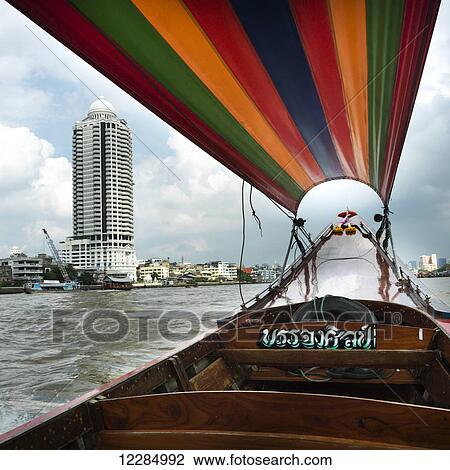Stock Image - Boat with colourful covering and buildings along the shoreline; Bangkok, Thailand. Fotosearch