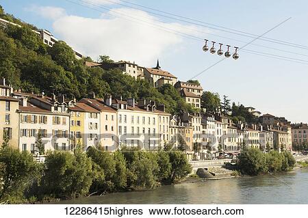 Stock Photograph - Bubble gondola passing over old buildings along a river; Grenoble, France. Fotosearch