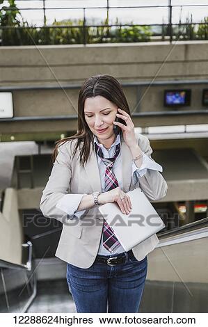 Stock Photograph - Business woman on an escalator talking on her cell phone, checking the time and holding a tablet; Edmonton, Alberta, Canada. Fotosearch