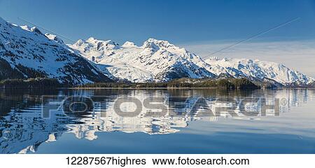 Stock Photograph - Calm ccean waters reflect snow covered mountains on a clear day in Kings Bay, Prince William Sound; Whittier, Alaska, United States of America. Fotosearch