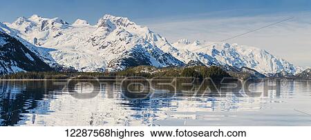 Stock Photograph - Calm ccean waters reflect snow covered mountains on a clear day in Kings Bay, Prince William Sound; Whittier, Alaska, United States of America. Fotosearch
