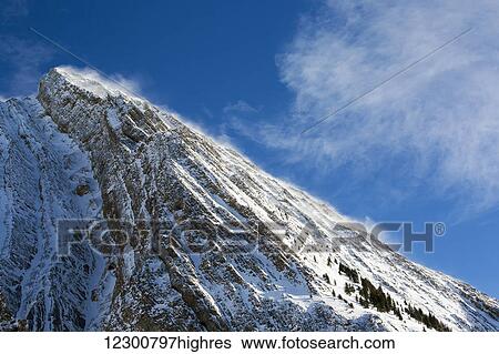 Close up of a snow covered mountain peak with wind blowing snow off the slope with blue sky and clouds; Kananaskis Country, Alberta, Canada View Large Photo Image Stock Photograph - Close up of a snow covered mountain peak with wind blowing snow off the slope with blue sky and clouds; Kananaskis Country, Alberta, Canada. Fotosearch