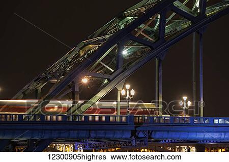 Stock Photograph - Close up of the illuminated arch of a bridge and lamp posts at nighttime. Fotosearch