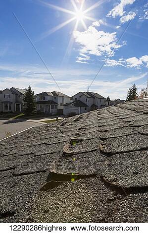 Stock Photograph - Close up of well worn rounded roof shingles with neighbourhood houses, sun burst, blue sky and clouds in the background; Calgary, Alberta, Canada. Fotosearch