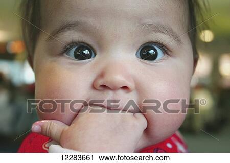 Stock Image - Close up of wide-eyed baby girl with her fingers in her mouth. her thumb wet and her eyes transfixed; Halifax, Nova Scotia, Canada. Fotosearch