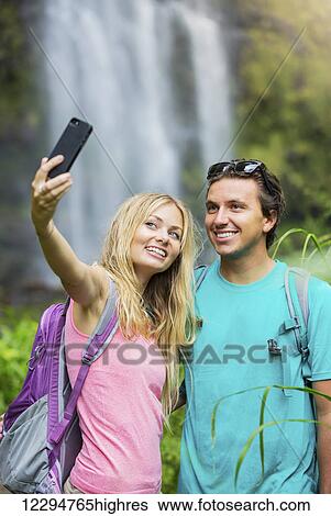 Stock Photograph - Couple having fun together outdoors. Taking self portrait with camera phone after hiking to incredible waterfall in Hawaii.. Fotosearch