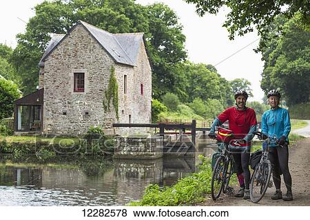 Stock Photo - Couple with bikes on a path along a canal with lock and stone lock house in the background; Gaec Grand Boutron, Calorguen, Brittany, France. Fotosearch