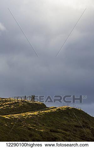 Stock Photograph - Dark cloudy sky with a rural road on a grassy slope; Lothian, Scotland. Fotosearch