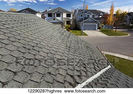 Stock Photograph - Detail of well worn rounded roof shingles with neighbourhood houses, autumn colours, blue sky and clouds in the background; Calgary, Alberta, Canada. Fotosearch