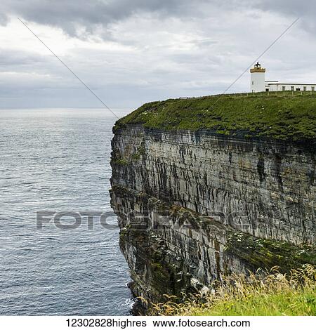 Stock Photograph - Duncansby Head Lighthouse on the edge of a cliff on the coast under a cloudy sky; Scotland. Fotosearch