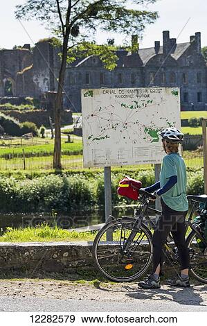Female cyclist looking at a large map sign along a canal with an Abbey in the background; Saint-Gelven, Brittany, France View Large Photo Image Stock Photo - Female cyclist looking at a large map sign along a canal with an Abbey in the background; Saint-Gelven, Brittany, France. Fotosearch