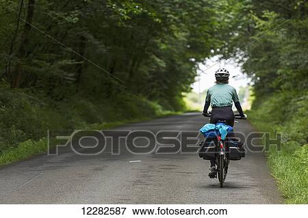 Stock Photo - Female cyclist on paved road with a canopy of trees creating a tunnel in the distance; Plousne, Brittany, France. Fotosearch