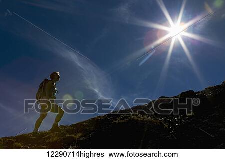 Stock Photograph - Female hiker climbing a rocky slope with blue sky and a sun burst; Waterton, Alberta, Canada. Fotosearch