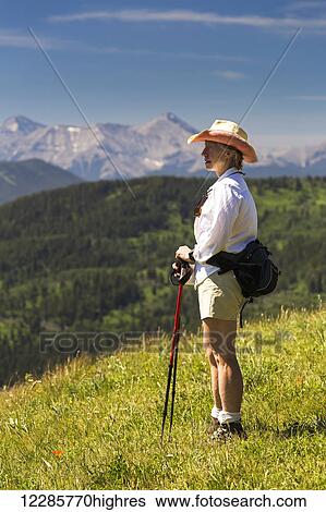 Stock Photograph - Female hiker on top of grassy hill overlooking rolling foothills with mountain range in the background with blue sky; Kananaskis Country, Alberta, Canada. Fotosearch