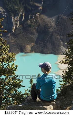 Stock Photograph - Female hiker sitting on rock high above intense blue coloured mountain lake with mountain cliffs in the background, Banff National Park; Alberta, Canada. Fotosearch