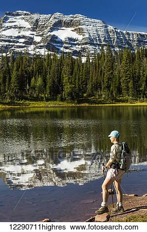 Stock Photograph - Female hiker standing on shoreline of mountain lake reflecting snow peaks and blue sky; Waterton, Alberta, Canada. Fotosearch