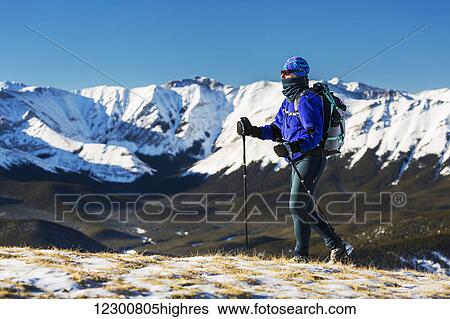 Stock Photograph - Female hiker walking on top of a snow covered ridge with foothills below and snow covered mountain range and blue sky in the background; Kananaskis Country, Alberta, Canada. Fotosearch