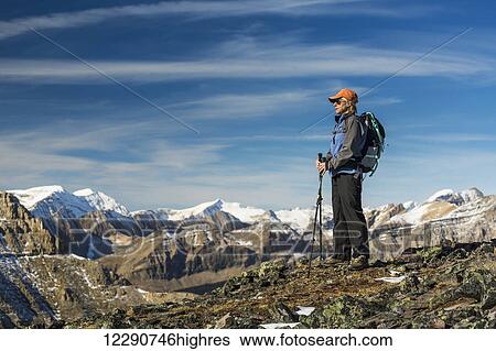 Female hiker with poles standing on top of a rocky mountain ridge overlooking snow peaked mountain ranges with blue sky and clouds, Banff National Park; Alberta, Canada View Large Photo Image Stock Photograph - Female hiker with poles standing on top of a rocky mountain ridge overlooking snow peaked mountain ranges with blue sky and clouds, Banff National Park; Alberta, Canada. Fotosearch