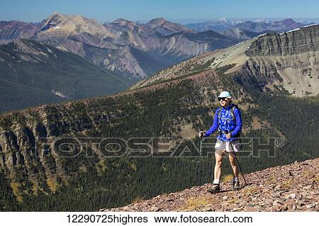 Stock Photograph - Female hiker with poles walking along a mountain ridge with mountain ranges and blue sky in the background; Waterton, Alberta, Canada. Fotosearch