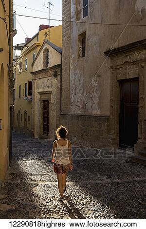 Stock Photograph - Girl walking down a cobblestone alley between buildings; Alghero, Sardinia, Italy. Fotosearch