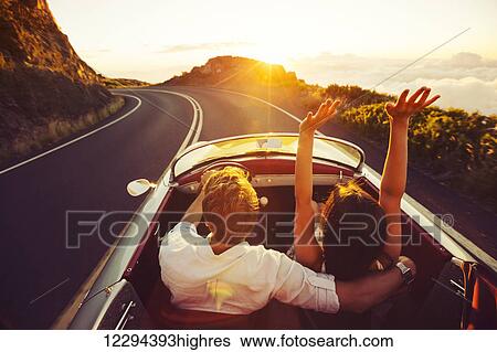 Stock Photograph - Happy Couple Driving on Country Road into the Sunset in Classic Vintage Sports Car. Fotosearch