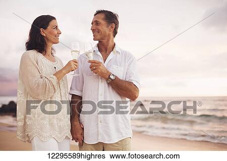 Stock Photograph - Happy Romantic Mature Couple Drinking Champagne on the Beach at Sunset. Vacation Travel Retirement Anniversary Celebration.. Fotosearch
