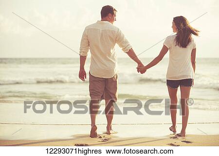 Stock Photograph - Happy Romantic Young Couple Walking Down the Beach at Sunset on Vacation. Fotosearch