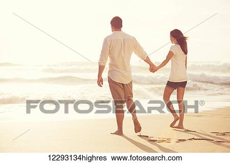 Stock Photograph - Happy Romantic Young Couple Walking Down the Beach at Sunset on Vacation. Fotosearch