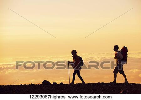 Stock Photograph - Hikers with backpacks hiking on top of a mountain at sunset. Fotosearch