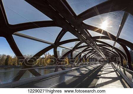 Stock Photograph - Interesting silhouette design of cylindrical bridge with sunburst and trees and buildings in the background; Calgary, Alberta, Canada. Fotosearch