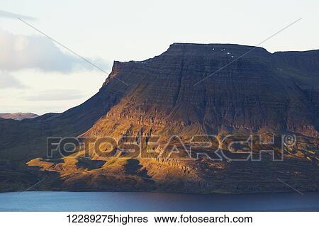 Stock Photograph - Late day sun illuminates the steep tiered cliffs in Iceland's Westfjords; Westfjords, Iceland. Fotosearch