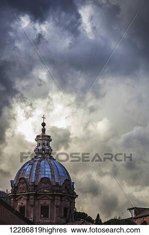 Stock Photograph - Light breaks through the turbulent clouds on the dome of the Santi Luca e Martina church; Rome, Italy. Fotosearch