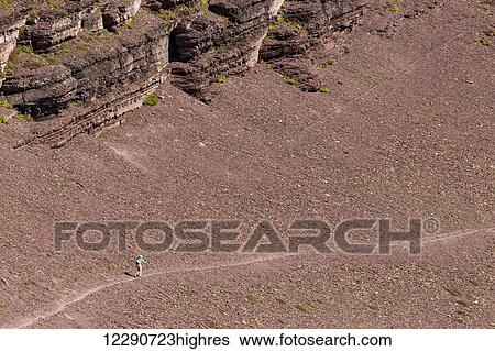 Stock Photograph - Lone female hiker on vast red rock slope of scree with mountain cliffs above; Waterton, Alberta, Canada. Fotosearch