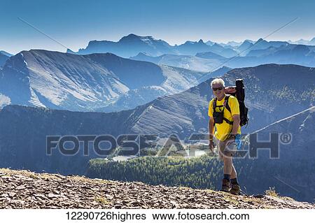 Stock Photograph - Male hiker ascending a rocky peak with mountain ranges and lakes in the background; Waterton, Alberta, Canada. Fotosearch