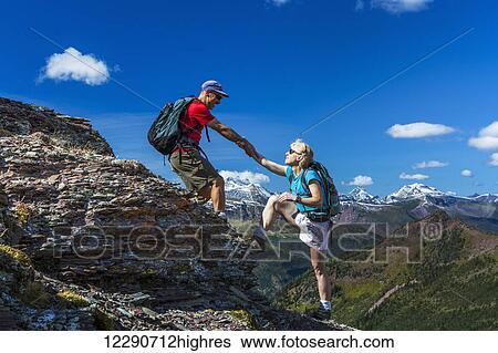 Stock Photograph - Male hiker helping female hiker up a rocky peak of a mountain ridge with grand vista and blue sky and clouds in the background; Waterton, Alberta, Canada. Fotosearch