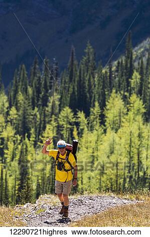 Male hiker on mountain trail waving with brightly lit trees and mountain cliff in the background; Waterton, Alberta, Canada View Large Photo Image Stock Photograph - Male hiker on mountain trail waving with brightly lit trees and mountain cliff in the background; Waterton, Alberta, Canada. Fotosearch