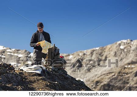 Stock Photograph - Male hiker on top of a mountain looking at map with mountain range and blue sky in the background, Kananaskis Provincial Park; Alberta, Canada. Fotosearch
