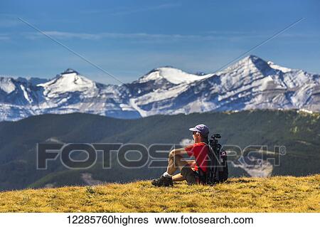 Stock Photograph - Male hiker sitting on a grassy mountain top overlooking foothills, snow capped mountain range and blue sky with clouds; Kananaskis Country, Alberta, Canada. Fotosearch