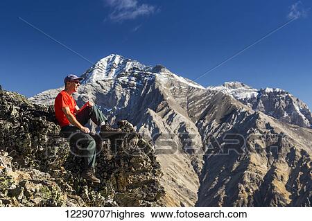 Male hiker sitting on top of mountain ridge overlooking snow peaked mountains in the background with blue sky and clouds, Kananaskis Provincial Park; Alberta, Canada View Large Photo Image Stock Photograph - Male hiker sitting on top of mountain ridge overlooking snow peaked mountains in the background with blue sky and clouds, Kananaskis Provincial Park; Alberta, Canada. Fotosearch