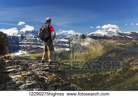 Male hiker standing on top of mountain ridge overlooking snow peaked mountains and forest valley below with blue sky and clouds; Waterton, Alberta, Canada View Large Photo Image Stock Photograph - Male hiker standing on top of mountain ridge overlooking snow peaked mountains and forest valley below with blue sky and clouds; Waterton, Alberta, Canada. Fotosearch