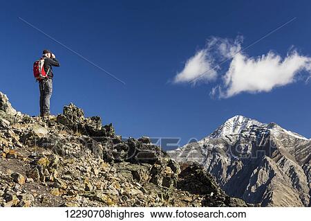 Stock Photograph - Male hiker standing on top of mountain ridge looking through binoculars at distant snow peaked mountain with blue sky and clouds, Kananaskis Provincial Park; Alberta, Canada. Fotosearch